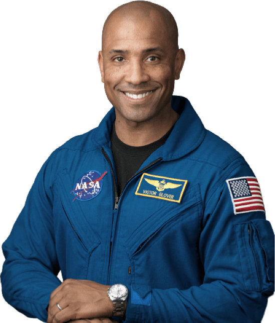 Pilot Victor Glover, a bald guy with a big friendly smile in a blue NASA overall with a black shirt under it and a silver watch, posing slightly sideways with one hand on the other.