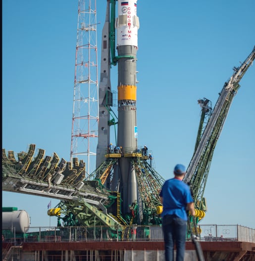 A spaceship rigged in the spaceport being prepared for launch by people in blue shirts.